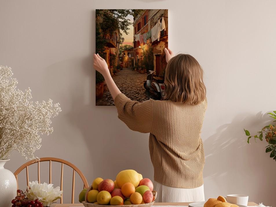A woman hangs a framed picture of a cobblestone street in a village on a wall in a cozy, homely setting.