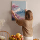 A woman hangs a framed picture of a mountain with pink flowers on a wall in a cozy room with a dining table, chairs, and various fruits.