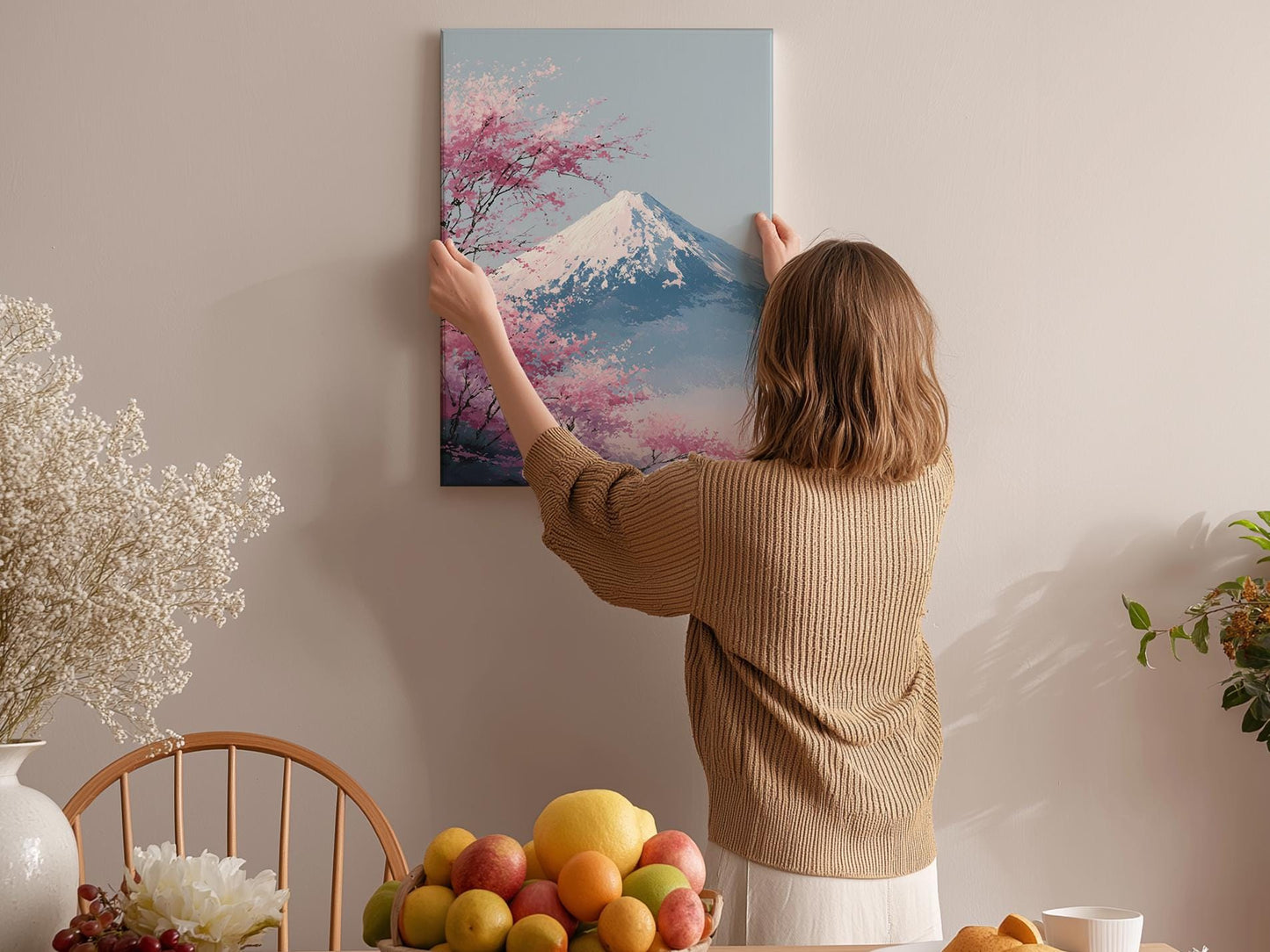 A woman hangs a framed picture of a mountain with pink flowers on a wall in a cozy room with a dining table, chairs, and various fruits.