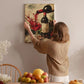 A woman hangs a painting of a wine glass and bottle on a wall in a cozy kitchen setting.