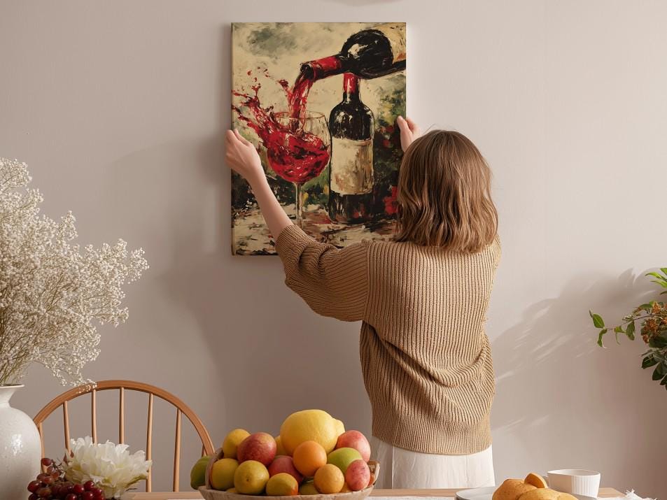 A woman hangs a painting of a wine glass and bottle on a wall in a cozy kitchen setting.