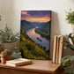 a wooden desk with a framed landscape photograph of a river valley, surrounded by mountains and a sunset sky. The photograph is placed on the desk, which also has several potted plants, books, and other decorative items.