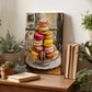 a wooden table with a painting of colorful macarons on a glass plate, surrounded by potted plants and books.