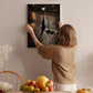 A woman hangs a framed photograph of a cobblestone street at night on a wall in a cozy, homely setting.