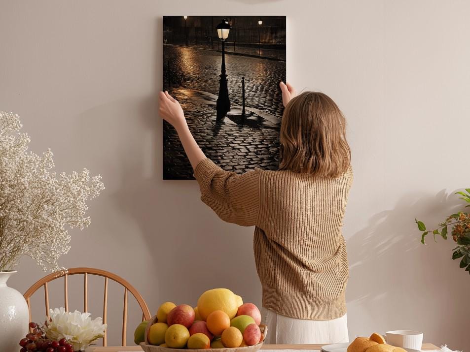 A woman hangs a framed photograph of a cobblestone street at night on a wall in a cozy, homely setting.